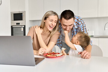 Obraz na płótnie Canvas Happy young family couple with kid son having fun eating toasts with jam using laptop in kitchen. Cute small child boy and parents enjoying breakfast snack while working from home on computer.
