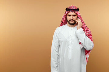 Arab man businessman in national clothes talking on a smartphone, like, beige background. Dishdasha, kandora, thobe, middle east traditional menswear concept, islam. Copy space.