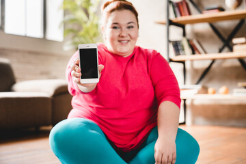 Fat obese caucasian plus size woman showing blank phone`s screen after hard physical training at home for losing weight and burning fat with calories on mat