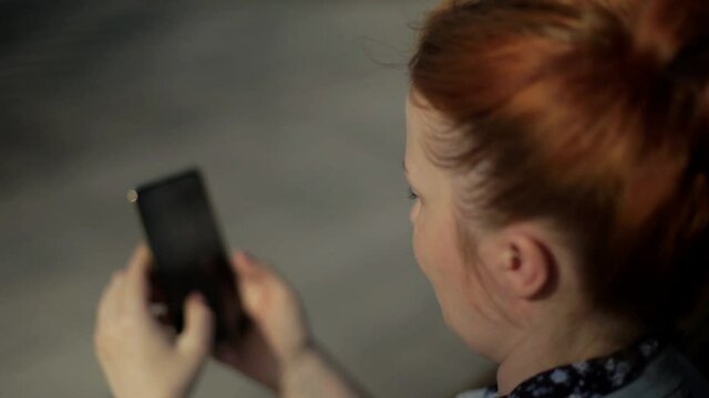 young red-haired woman with a phone on a bench in the park, communicating on social networks using a mobile device, live camera effect