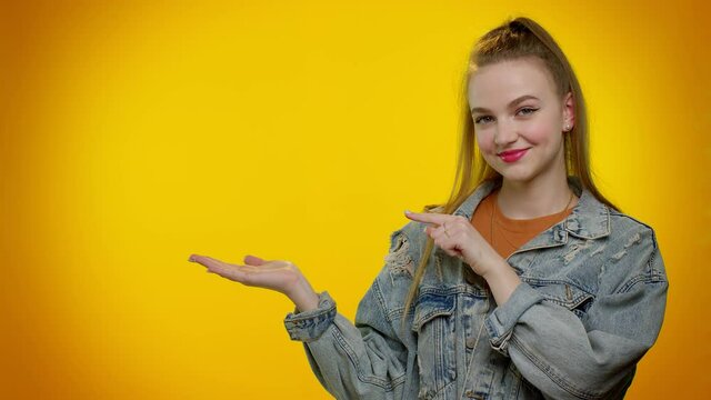 Pretty stylish teenager girl showing thumbs up and pointing empty place, advertising area for commercial text, copy space for goods promotion. Young adult woman. Studio shot on yellow wall background