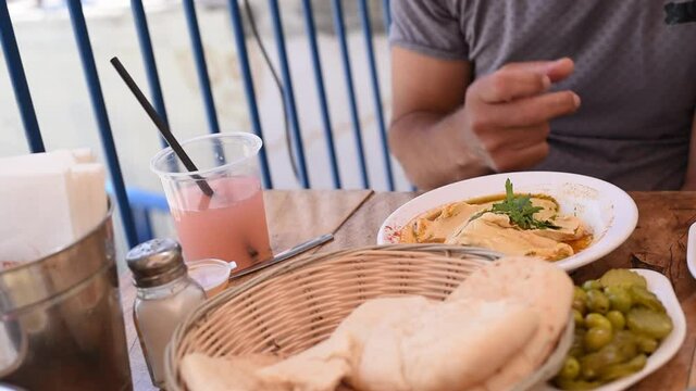 Man's hand takes piece pita and dunk in hummus. Pita bread in wicker basket, hummus with oil and parsley, olives and pickles on white plates on wooden table. Arabic traditional cuisine. Close-up.