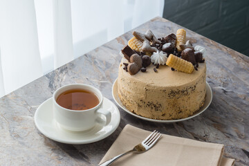 A cup of tea on a saucer and a sponge cake with buttercream on marble countertop. The cake is decorated with chocolate and cookies