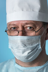 Portrait of middle-aged experienced doctor in a medical cap and mask, wearing glasses. Close-up view of the camera