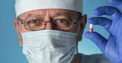 An elderly doctor in medical clothing: cap, gloves,dressing gown and mask, glasses. He holds a medicine capsule in his hands . Close-up, look at the camera