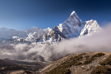A mountain landscape in the Himalayas featuring mount Ama Dablam against vivid blue sky with low clouds going from the right and a hill grown with sparse bushes in the foreground
