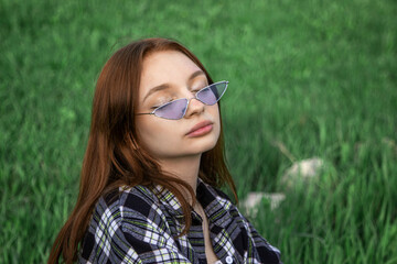 Portrait of a teenage girl on a background of grass. Young girl in triangular glasses. Girl with red hair