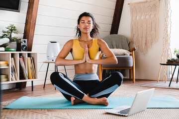 Serene young fit african woman meditating at home using laptop. Home Training. Relaxed woman in practicing online sports, watching video tutorial on laptop computer, doing stretching yoga exercises