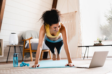 Fit sporty african-american young woman standing in plank position using laptop at home. Cheerful African woman stretching up In front of laptop, doing home workout,