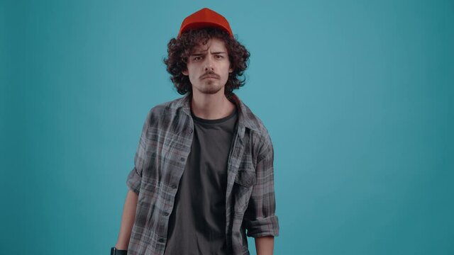 The charismatic young man, energetically puts his cap on his head, looks questioningly forward and then angrily jumps up and breathes his sleeve. isolated over turquoise background.Concept of life