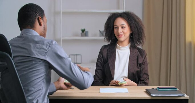 Unrecognizable afro american business man sitting at office table gives money salary dollars to confident curly woman specialist consultant lawyer accountant pays for services shaking female hand - Powered by Adobe