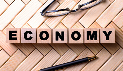 The word ECONOMY is written on wooden cubes on a wooden background next to a pen and glasses.