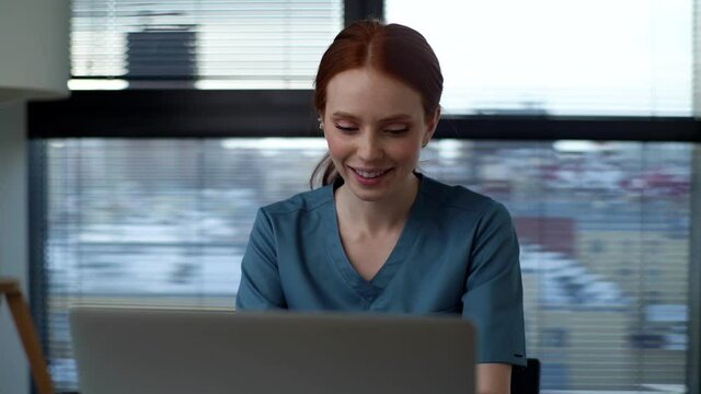Front view of smiling young female doctor in blue green medical uniform typing on laptop keyboard sitting at desk on background of window in modern office of medic clinic. Tracking shot in slow motion