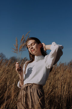 Smiling young woman in white blouse standing in field and holding braches of dry pampas grass in front of sky. Style and fashion. Girl in casual outfit with closed eyes touching hair. Golden hour
