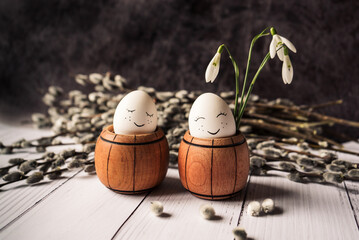 white eggs with a cute smile in a small wooden barrel with snowdrops and pussy willow branches with seals on a gray background