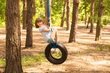 Cute little girl swinging on wheel attached to big tree in autumn forest. 