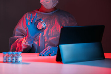 Vaccine approval. Covid-19 telehealth. Virtual care. Medical advice online. Female doctor in ppe gloves showing vial dose on tablet online in red blue neon light in dark laboratory.