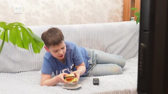 a teenage boy at home lies on the couch and watches TV, cartoons or movies, switches channels, programs. Eating a hamburger. Free time. Leisure. modern youth