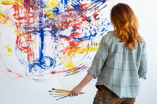 Art therapy. Hand painting. Creative hobby. Back view portrait of talented redhead woman artist with paintbrushes looking at blur colorful blue red yellow abstract picture on white wall.