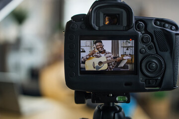 Cheerful young man with dark skin recording video on camera while playing guitar. Happy male blogger creating music content for his social networks.
