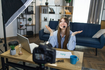 Charming caucasian woman in headset making live broadcast using modern video camera. Pretty female...