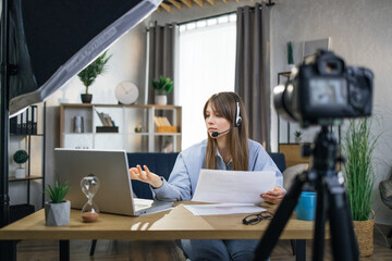 Qualified teacher conducting online lessons using headset, laptop and modern camera. Pretty woman sitting at table, talking and gesturing on camera.