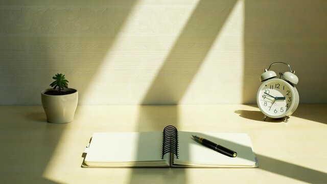 Shadow of an alarm clock, a notebook, a pen and a green plant on desk under sun