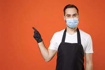 Young man barista barman employee in apron white t-shirt face mask coronavirus covid19 pandemic quarantine work coffee shop point finger aside workspace isolated on orange background Business startup