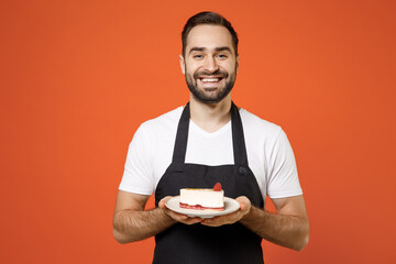 Young man 20s barista bartender barman employee in black apron white t-shirt work coffee shop holding sweet cake dessert isolated on orange background studio portrait Small business startup concept.