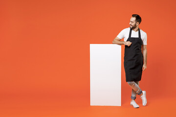 Full length young man barista bartender barman employee in apron t-shirt work in coffee shop leaning on blank board promotional content place text isolated on orange background Small business startup.