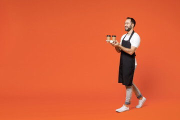 Full length young fun man barista barman employee in black apron white t-shirt work in coffee shop hold paper takeaway delivery cups walk go isolated on orange background Small business startup. - obrazy, fototapety, plakaty