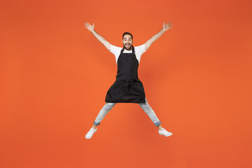 Fototapeta na wymiar Full length young man barista bartender barman employee in black apron white tshirt work in coffee shop jump high outstretched hands legs isolated on orange background. Small business startup concept