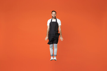 Fototapeta na wymiar Full length young fun man 20s barista bartender barman employee in black apron white t-shirt work in coffee shop jump high fly gesture isolated on orange background. Small business startup concept.