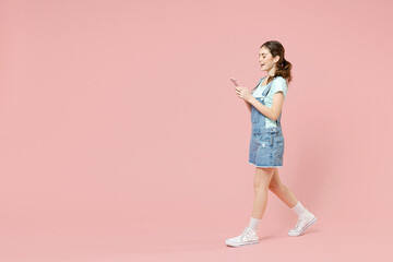 Full length side view young smiling caucasian woman 20s wear trendy denim clothes blue t-shirt hold mobile cell phone walk isolated on pastel pink background studio portrait. People lifestyle concept