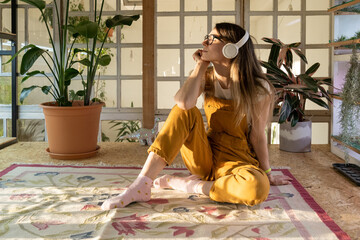 Satisfied woman wearing headset listen to music relaxed after remote work day at home. Cute female gardener sit on floor with closed eyes dreaming. Young casual girl small business owner relaxation