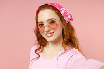 Close up fun young smiling happy caucasian woman 20s wearing rose clothes bandana glasses doing selfie shot on mobile phone isolated on pastel pink background studio portrait People lifestyle concept.