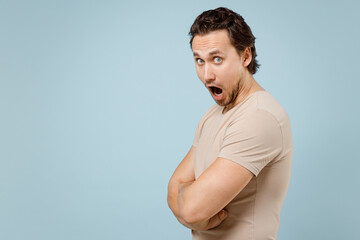 Side view young sad shocked indignant caucasian man wearing casual basic beige t-shirt looking camera hold hands crossed folded isolated on pastel blue background studio portrait. LIfestyle concept