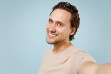 Close up young smiling happy fun attractive man 20s wearing casual basic beige t-shirt doing selfie shot on mobile phone isolated on pastel blue background studio portrait. People lifestyle concept.