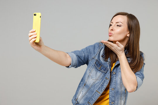 Young romantic girlfriend woman 20s wear in trendy denim jacket yellow t-shirt doing selfie shot on mobile phone post photo on social network blowing air kiss isolated on grey color background studio.