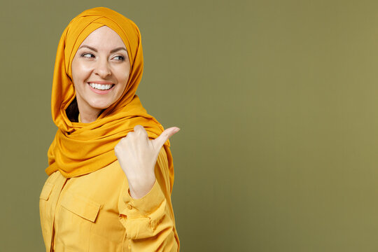 Smiling young arabian asian muslim woman in abaya hijab yellow clothes point thumb finger aside on workspace area isolated on olive green background People uae middle eastern islam religious concept.