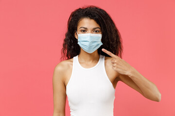 Young african american woman 20s wearing white tank shirt point index finger on sterile face mask ppe to safe from coronavirus virus covid-19 flu on lockdown quarantine isolated on pink background