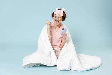 Young happy woman in pajamas jam sleep eye mask rest at home sit wrap cover under blanket duvet use mobile cell phone winner gesture isolated on pastel blue background Good mood night bedtime concept.