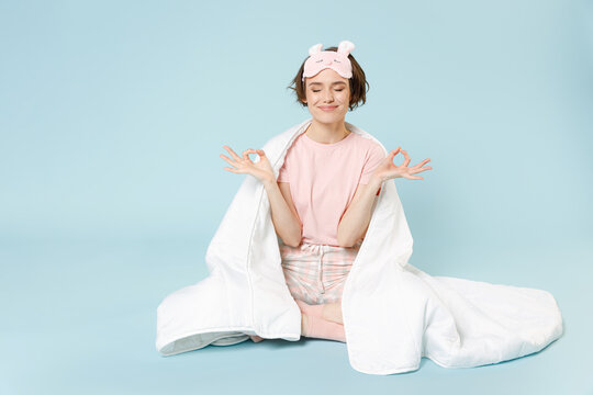 Young Woman In Pajamas Jam Sleep Eye Mask Rest Relax At Home Sitting Wrap Covered Under Blanket Duvet Do Yoga Om Gesture Calm Down Isolated On Pastel Blue Background Good Mood Night Bedtime Concept.