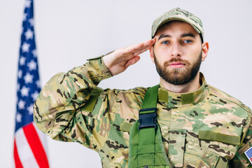 of handsome soldier in a military uniform salutes and looks to the camera near the American flag in studio on white background