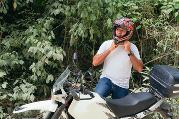 Naklejka premium Young man in white shirt and jeans strapping on a helmet on a motorcycle in the open air.