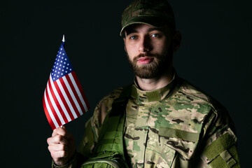Serious soldier in a military uniform holds American flag looks to the camera confident and serious in studio on dark background