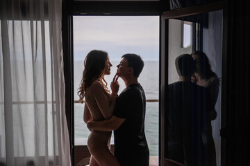 Silhouettes of young loving couple in front of panoramic window overlooking the sea. Lovers travel on their honeymoon, spend a date in hotel room. Beautiful woman hugs a man, kiss, look at each other