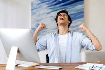 Joyful successful young Hispanic man, businessman, manager or freelancer, in stylish clothes, working at the computer, happy with victory, win or big profit, gesturing with his hands, shouting happily