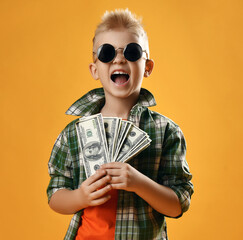 Blond smiling boy child in checkered shirt and sunglasses standing holding heap of money cash in hands feeling happy over yellow background. Trendy casual children fashion, business concept