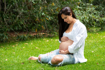 Naklejka na ściany i meble Beautiful portrait of pregnant woman sitting on the grass in a natural park. Maternity concept. Mother's day.